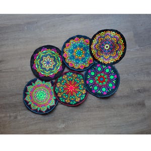 May include: Six round, black coasters with colorful mandala designs. Each coaster features a unique pattern in vibrant colors like green, pink, orange, and blue. The coasters are arranged on a wooden surface.