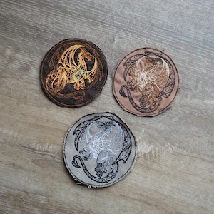 Op de afbeelding: Drie ronde geborduurde patches met drakenontwerpen. Eén is bruin met een gouden draak, een andere is beige met een bruine draak, en de derde is grijs met een witte draak. De patches liggen op een houten oppervlak.