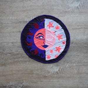 May include: A round, embroidered patch featuring a sun and moon design. The sun is on the left side and is orange with a purple outline. The moon is on the right side and is white with a pink outline. Both the sun and moon have a face with a single eye.