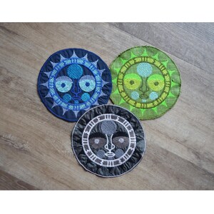 May include: Three embroidered patches featuring stylized faces with a sun-like design. The patches are circular and feature different color schemes: blue, green, and gray.
