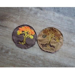 May include: Two round embroidered patches featuring stylized trees. One patch is brown with an orange and yellow tree design. The other patch is tan with a brown and yellow tree design. Both patches have a raw edge.