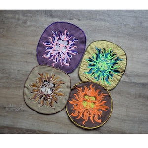 May include: Five embroidered patches with a sun face design. The patches are in various colours, including purple, tan, olive green, and brown, with the sun faces in shades of orange, yellow, blue, and pink. The patches are oval or round.