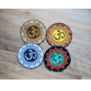 May include: Four round coasters with an Om symbol in the center. The coasters feature intricate mandala-style designs in brown, yellow, blue, and green, with a variety of background colors. The coasters are placed on a wooden surface.