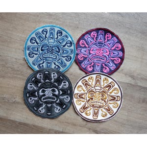 Op de afbeelding: Vier ronde geborduurde patches met een gestileerd zonontwerp met een gezicht. De patches zijn in blauw, roze, zwart en bruin, elk met een andere gekleurde rand. Het ontwerp heeft een Native American of tribale kunststijl.