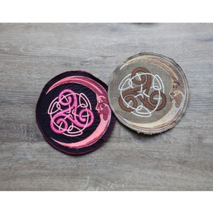 以下が含まれることがあります： 2つの円形刺繍パッチ。1つは黒で、三日月の中にピンクのケルトノットのデザインがあります。もう1つはタンで、三日月の中に茶色のケルトノットのデザインがあります。どちらも月に顔があります。