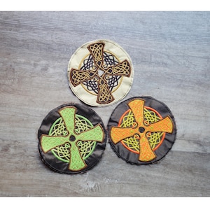 May include: Three embroidered Celtic cross patches. One is cream-coloured with brown and black details, another is black with green, and the third is black with orange and yellow. Each patch is round with a detailed cross design.