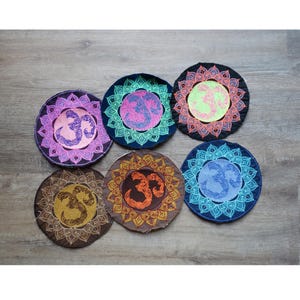Può includere: Sei sottobicchieri rotondi ricamati, ciascuno con un simbolo Om al centro. Ogni sottobicchiere presenta una diversa combinazione di colori, tra cui viola, verde acqua, nero, marrone, arancione e blu. Il simbolo Om è in un colore a contrasto e i sottobicchieri hanno un bordo decorativo.