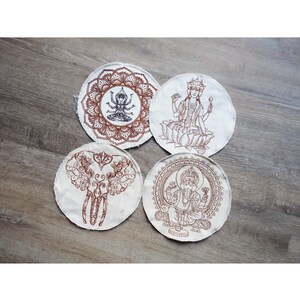 May include: Four round, off-white coasters with brown embroidered designs. The designs include a mandala, a deity seated on a lotus flower, an elephant, and a deity in a circular frame. The coasters are arranged on a wooden surface.