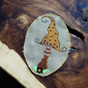 May include: Embroidered mushroom patch with a whimsical design. The mushroom features a yellow cap with brown spots, a striped stem, and a brown base. The patch is on a light green background with a yellow border. The patch is an oval shape.