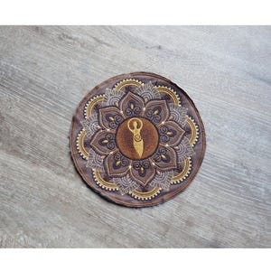 May include: Round, brown coaster with an embroidered mandala design. The coaster features a central figure and intricate floral patterns in shades of brown and gold. The coaster is made of fabric.