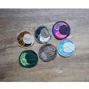 May include: A collection of six round embroidered patches featuring crescent moon designs. The patches showcase various color schemes, including brown and gold, black and white, purple and pink, blue and white, green and yellow, and gray and black. The patches are arranged on a wooden surface.