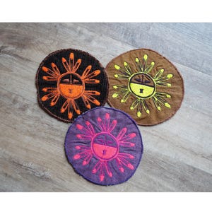 Könnte beinhalten: Drei gestickte Filzpatches mit einem Sonnendesign. Die Sonne hat ein Gesicht und Strahlen. Die Patches sind braun, lila und schwarz mit orange, gelben und rosa Strahlen.