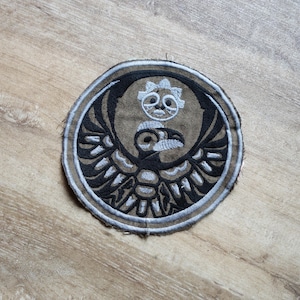 Può includere: Toppa rotonda ricamata con un disegno stilizzato di uccello in nero, bianco e marrone. La toppa ha una forma circolare con un emblema di uccello dettagliato al centro, circondato da un bordo.