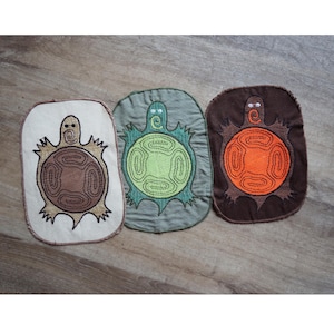 Op de afbeelding: Drie geborduurde schildpad-patches in verschillende kleuren. De schildpadden zijn bruin, groen en oranje. De patches bevinden zich op een houten oppervlak.