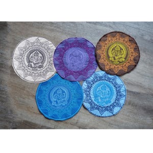 May include: Five circular fabric mats featuring embroidered Ganesha designs. The mats are white, purple, brown, light blue, and medium blue. Each mat displays a central Ganesha figure surrounded by intricate, radiating mandala-style patterns.