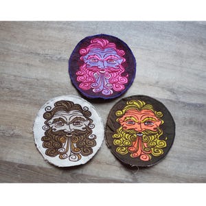 May include: Three round embroidered patches featuring a stylized bearded face. One patch has a purple background with pink and blue facial details. Another has a brown background with orange and yellow details. The third has a white background with brown details.
