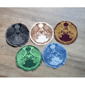 Puede incluir: Cinco parches redondos bordados con un diseño de nudo celta con aves. Los parches son de varios colores: negro, marrón, blanco, dorado, verde y azul. Los parches están sobre una superficie de madera.