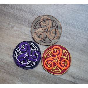 以下が含まれることがあります： ケルトノットのデザインが施された、3つの刺繍パッチ。1つはタンとブラウン、もう1つは黒い背景に紫と白、3つ目は赤とオレンジです。パッチは円形で、ステッチされた縁取りがあります。
