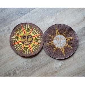 Puede incluir: Dos parches bordados que representan soles con caras. Un sol es amarillo con una cara roja y el otro es blanco con una cara marrón. Ambos soles tienen rayos marrones.