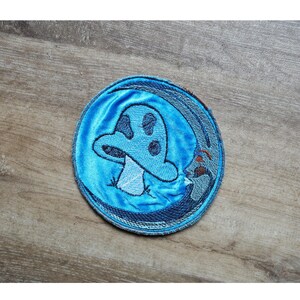 Op de afbeelding: Een ronde, blauw geborduurde patch met een gestileerd paddestoelontwerp. De paddestoel is lichtblauw met donkerblauwe stippen en een witte steel. De patch heeft een denim-achtige rand en een fluwelen midden.