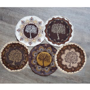 May include: Five circular fabric coasters featuring embroidered tree designs. Each coaster displays a central tree motif surrounded by intricate, decorative border patterns in shades of brown, cream, gold, and dark tan, arranged in a cluster.