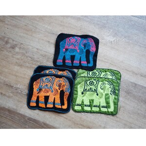 May include: Embroidered elephant coasters in blue, orange, and green. Each coaster features a detailed elephant design with intricate patterns. The coasters are square-shaped and set against a wooden background.