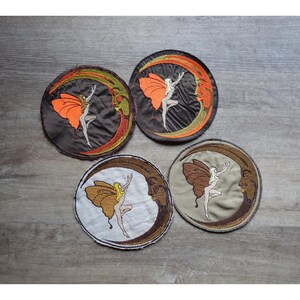 Peut inclure: Quatre écussons ronds brodés représentant une fée dansante avec des ailes de papillon et un croissant de lune. Les écussons présentent des couleurs orange, marron et beige, avec un design détaillé et une bordure cousue.