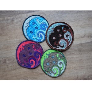 May include: Four round embroidered patches with celestial designs. Each patch features a crescent moon, stars, and swirling patterns in various colors: blue, brown, purple, and green. The patches are on a wooden surface.