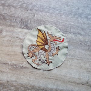 May include: Embroidered patch featuring a brown dragon with tan wings and a red tongue, set against a light green circular background. The dragon is detailed with white and brown accents, and the patch has a stitched edge.