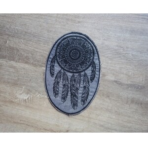 May include: Oval-shaped, gray patch featuring a black dreamcatcher design. The dreamcatcher includes a central mandala pattern and hanging feathers. The patch has a distressed, frayed edge, giving it a vintage look.