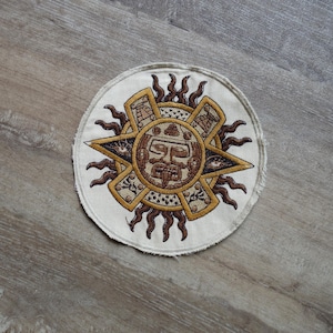 Könnte beinhalten: Runder, gestickter Aufnäher mit einem detaillierten aztekischen Sonnendesign in Braun-, Beige- und Goldtönen. Der Aufnäher hat einen cremefarbenen Hintergrund und eine dekorative Bordüre. Das Sonnendesign umfasst ein zentrales Gesicht und strahlende Flammen.