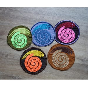 Könnte beinhalten: Fünf runde, gestickte Aufnäher mit einem stilisierten Drachen- und Spiralmuster. Die Aufnäher sind in verschiedenen Farben gehalten: Grün, Blau, Rosa, Orange und Braun. Sie sind auf einer Holzoberfläche platziert, jedes mit einem einzigartigen Design.