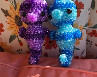May include: Two crocheted seahorse figurines, one purple and one blue, with black eyes and a pink nose. The seahorses are sitting on a pink surface.