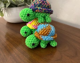 May include: Two green rubber band turtles stacked on top of each other. The top turtle has a multi-colored shell and the bottom turtle has a blue, gold, and brown shell.