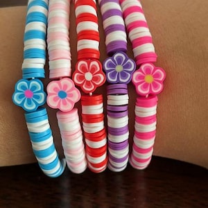 May include: Five colorful bracelets with flower charms. Each bracelet is made of stacked, round beads in alternating colors of white and blue, pink and white, red and white, purple and white, and pink and white. The flower charms are blue, pink, red, purple, and yellow.