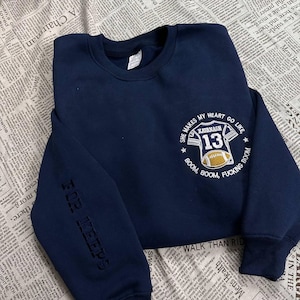 Puede incluir: Sudadera azul marino con el texto "SHE MAKES MY HEART GO LIKE" y un gráfico de camiseta de fútbol. La camiseta tiene el nombre "KAVANAGH" y el número "13". La manga dice "FOR KEEPS".