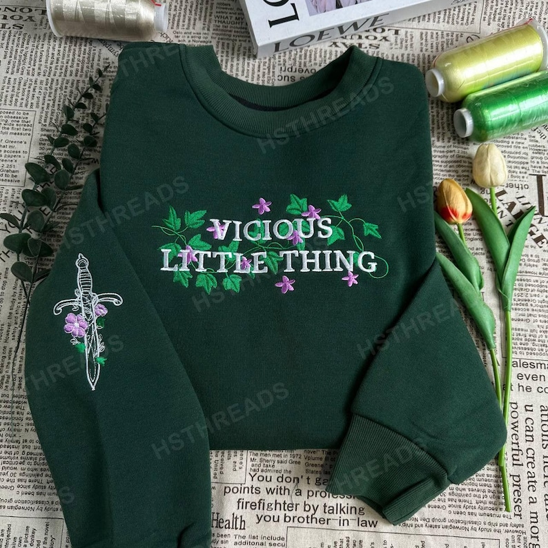 Pu&ograve; includere: Una felpa verde scuro con un design ricamato bianco che dice "Vicious Little Thing" con un pugnale e fiori.