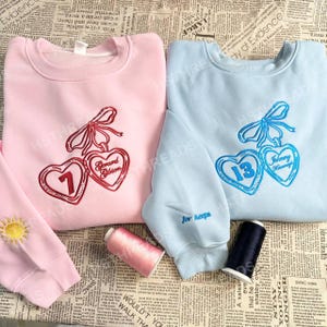 Peut inclure: Deux sweatshirts, un rose et un bleu clair, avec des motifs de cœurs brodés. Le sweat-shirt rose a un motif de cœur rouge avec le texte "Gerard Gibson" et le chiffre "1". Le sweat-shirt bleu a un motif de cœur bleu avec le texte "Johnny Kamang" et le chiffre "13".