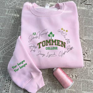 Puede incluir: Una sudadera rosa claro con el texto "Tommen College" en verde y blanco, junto con firmas y un trébol. La manga dice "For keeps" en verde. También se ve un carrete de hilo rosa.
