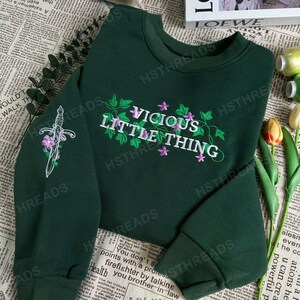 Pu&ograve; includere: Una felpa verde scuro con il testo "Vicious Little Thing" ricamato in bianco e viola. La felpa presenta un design floreale con viti e fiori. La felpa ha un pugnale ricamato sulla manica sinistra.