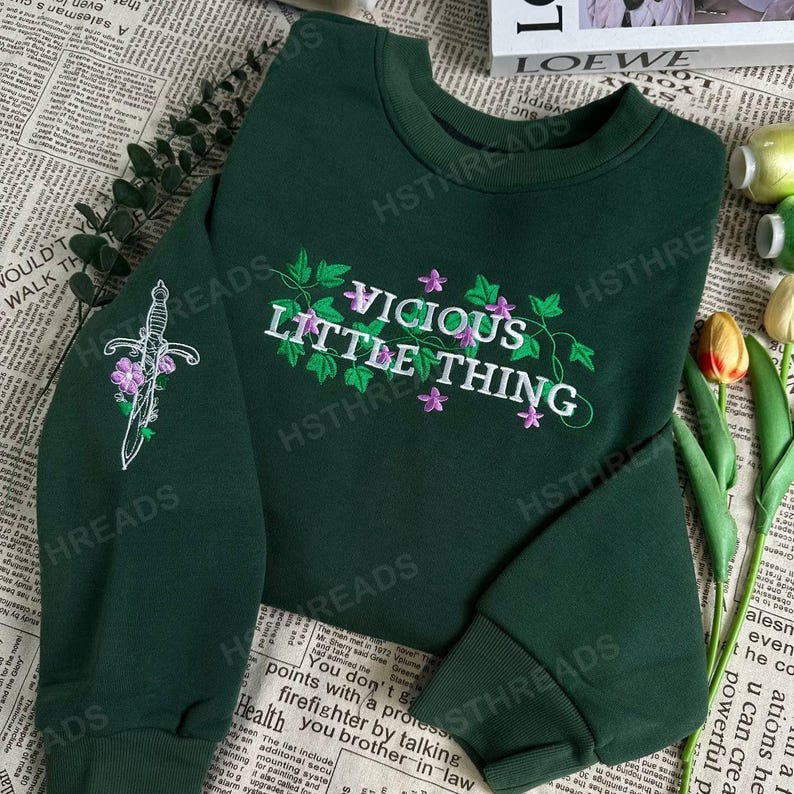 Pu&ograve; includere: Una felpa verde scuro con il testo "Vicious Little Thing" ricamato in bianco e viola. La felpa presenta un design floreale con viti e fiori. La felpa ha un pugnale ricamato sulla manica sinistra.