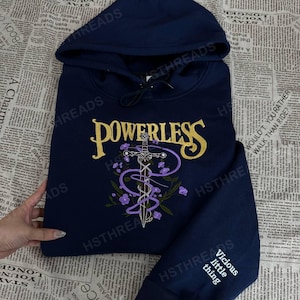 Könnte beinhalten: Marineblauer Kapuzenpullover mit dem Wort "POWERLESS" in goldener Schrift über einem Schwertdesign mit lila Akzenten. Der Ärmel zeigt den Text "Vicious little thing" in Weiß.