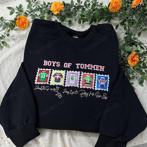 May include: Black sweatshirt with the text "BOYS OF TOMMEN" in pink. Below the text are five colorful stamp-like designs featuring jersey numbers and signatures. The sweatshirt is a crew neck style.