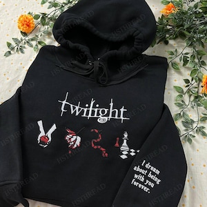 Puede incluir: Sudadera con capucha negra con la palabra "Twilight" bordada en blanco. El diseño incluye imágenes bordadas de una manzana roja, una rosa, una serpiente y una pieza de ajedrez. La manga tiene el texto "I dream about being with you forever."