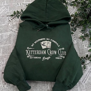 Pode incluir: Uma camisola com capuz verde floresta escuro com texto bordado a branco que diz No Mourners No Funerals, Ketterdam Crow Club e Dregs, juntamente com uma ilustração gráfica de três cartas de jogar com um corvo e uma espada.