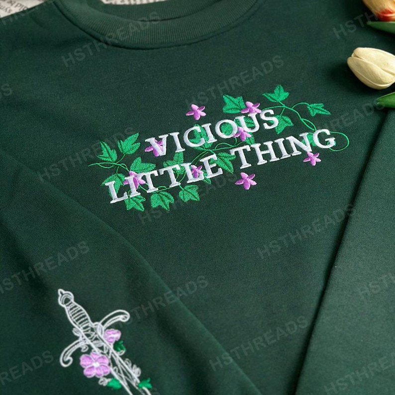 Pu&ograve; includere: Una felpa verde scuro con un ricamo bianco che dice "Vicious Little Thing" con foglie verdi e fiori rosa.