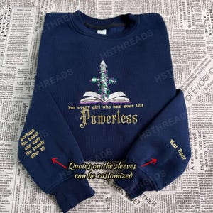 Op de afbeelding: Een donkerblauw sweatshirt met een gouden geborduurd ontwerp van een boek, een zwaard en bloemen. De tekst "For every girl who has ever felt Powerless" is geborduurd op het sweatshirt. De mouwen hebben de tekst "Quotes on the sleeves can be customized" en "Kai Azer" geborduurd.
