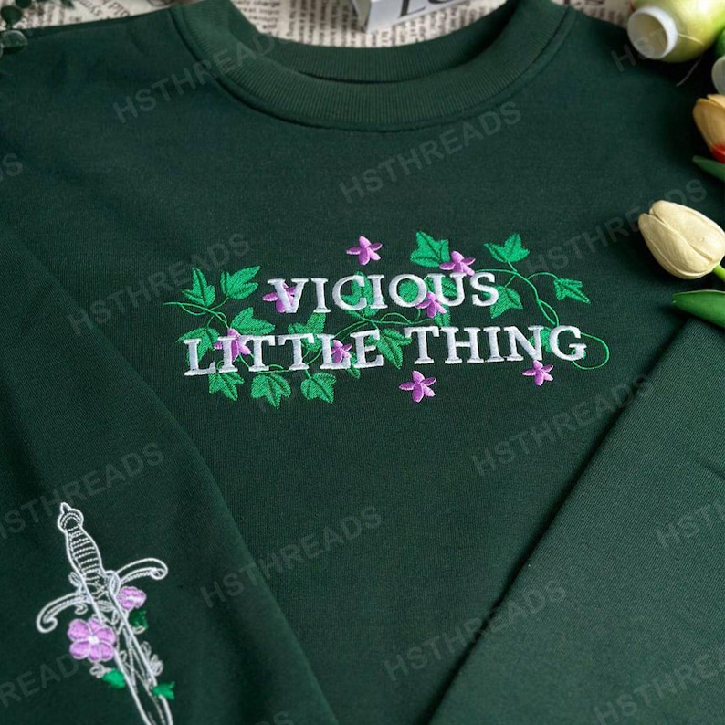 Pu&ograve; includere: Una felpa verde scuro con il testo "VICIOUS LITTLE THING" ricamato in bianco con fiori viola e foglie verdi.