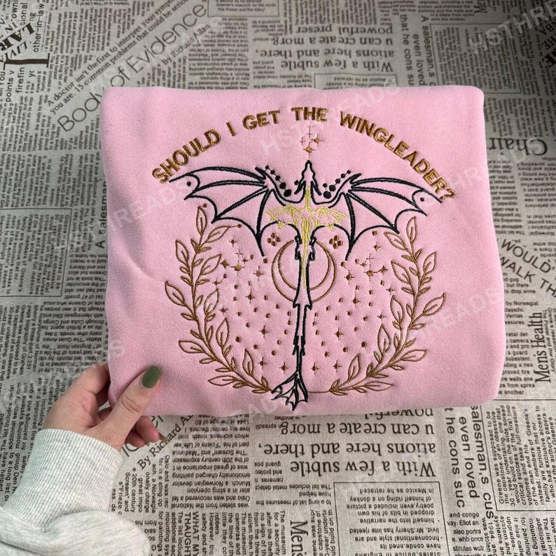 May include: Pink sweatshirt with embroidered text that reads "Should I get the Wingleader?" and a black and gold dragon with wings spread.