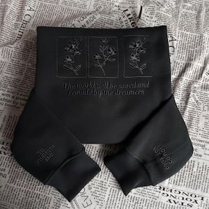 Puede incluir: Una sudadera negra de manga larga con tres paneles florales bordados en el pecho. Debajo de las flores, el texto dice: "The world will be saved and remade by the dreamers". El puño izquierdo dice "TO WHATEVER END" y el derecho "YOU DO NOT YIELD".